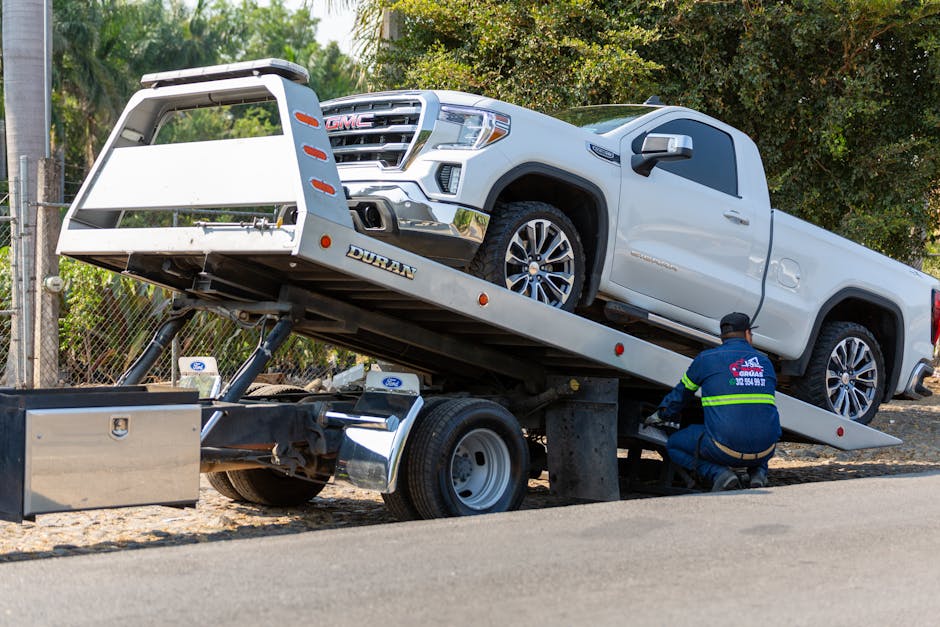 Tow truck operator in a hi-vis safety vest preparing a flatbed tow truck to load a white GMC pickup