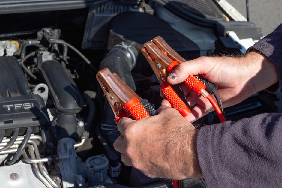 Hands holding orange jumper cable clamps over a car engine bay during a battery jumpstart